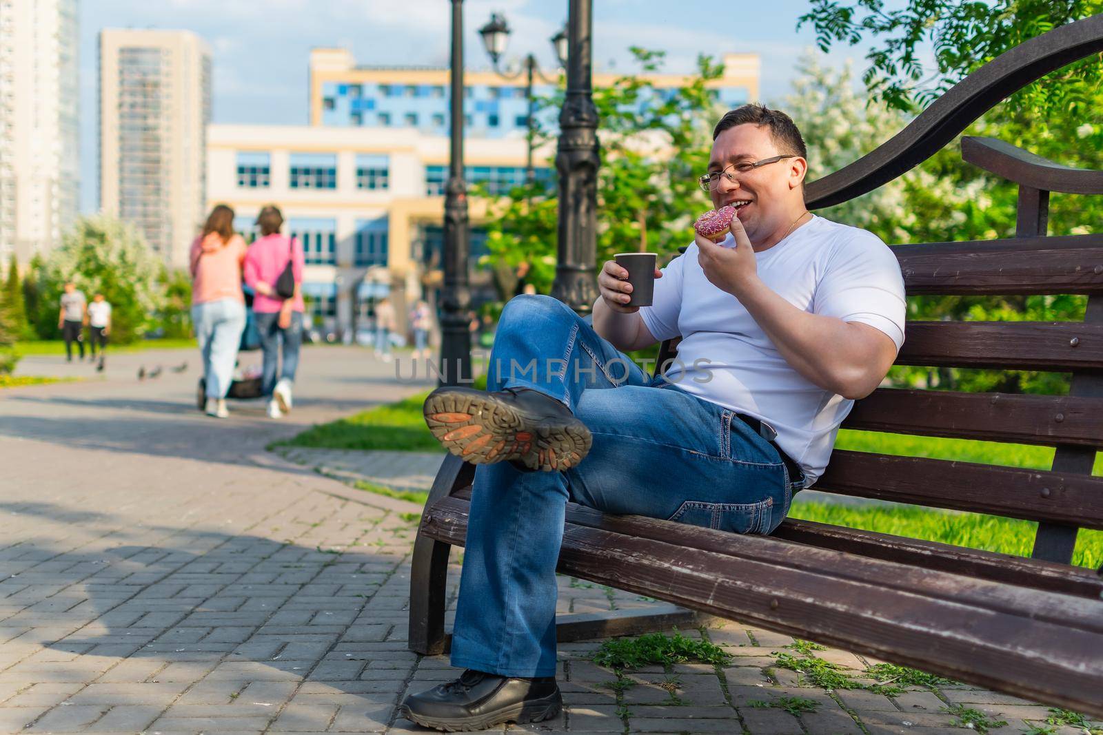 Happy man enjoying delicious donut and drink, laughing while sitting on a park bench