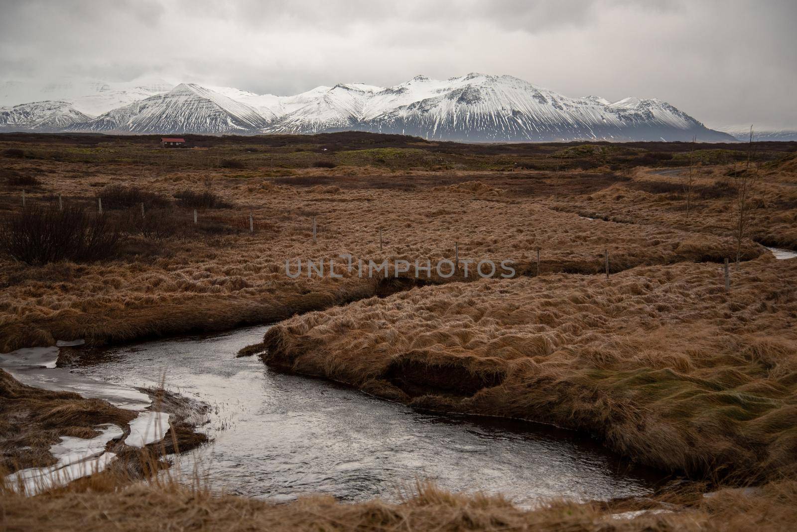 Icelandic landscape with stream through grassy field with snow capped mountains in the distance atmospheric cloudy textured by jyurinko