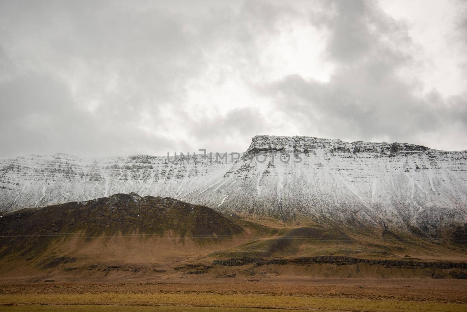 Heavenly light through the clouds over a snow capped mountain ridge leading down into brown grassy landscape in Iceland by jyurinko