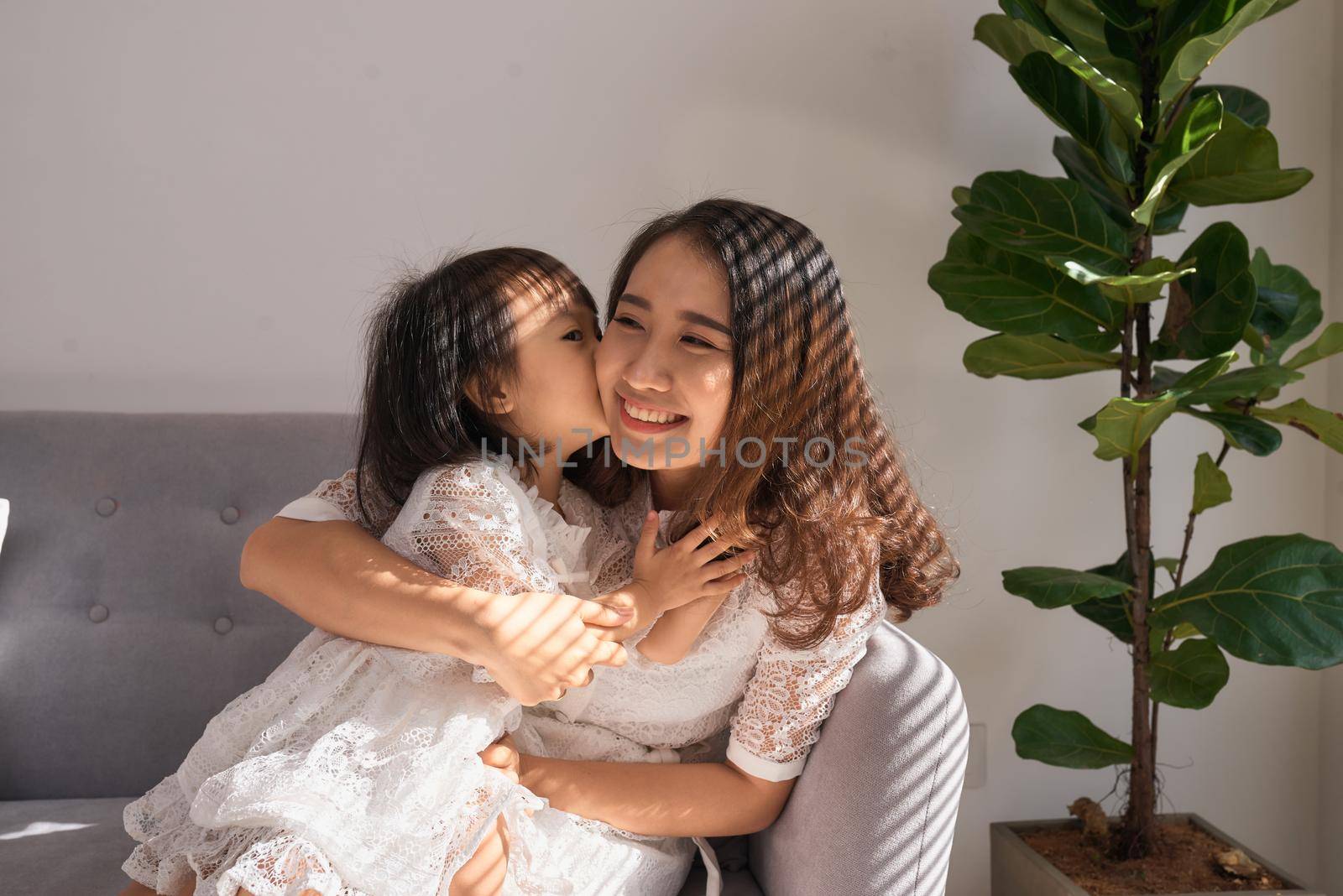 Sweet little girl is hugging and kissing her beautiful young mom in cheek while sitting on couch at home by makidotvn