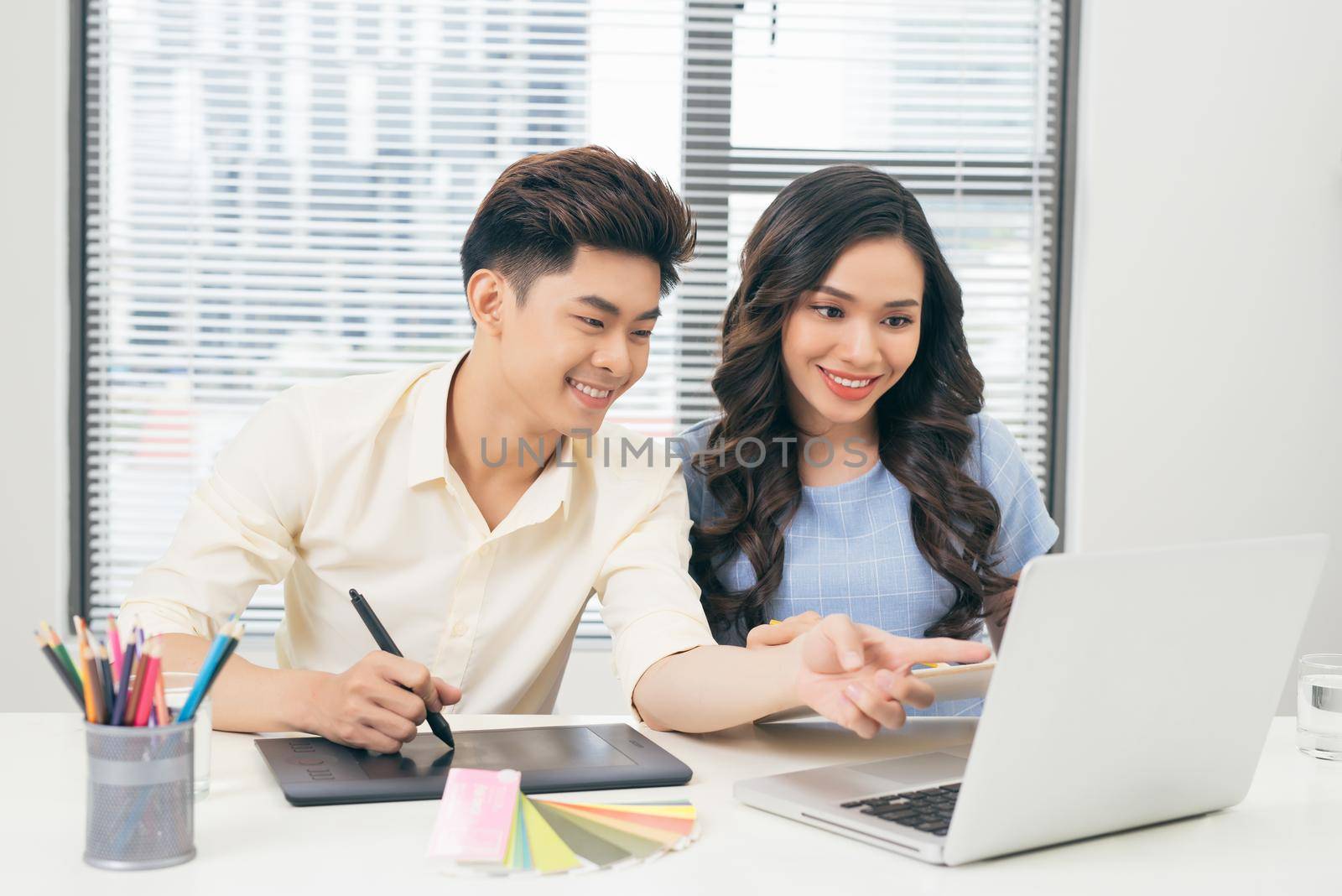 Two smiling casual designers working with laptop while sitting at desk in the office