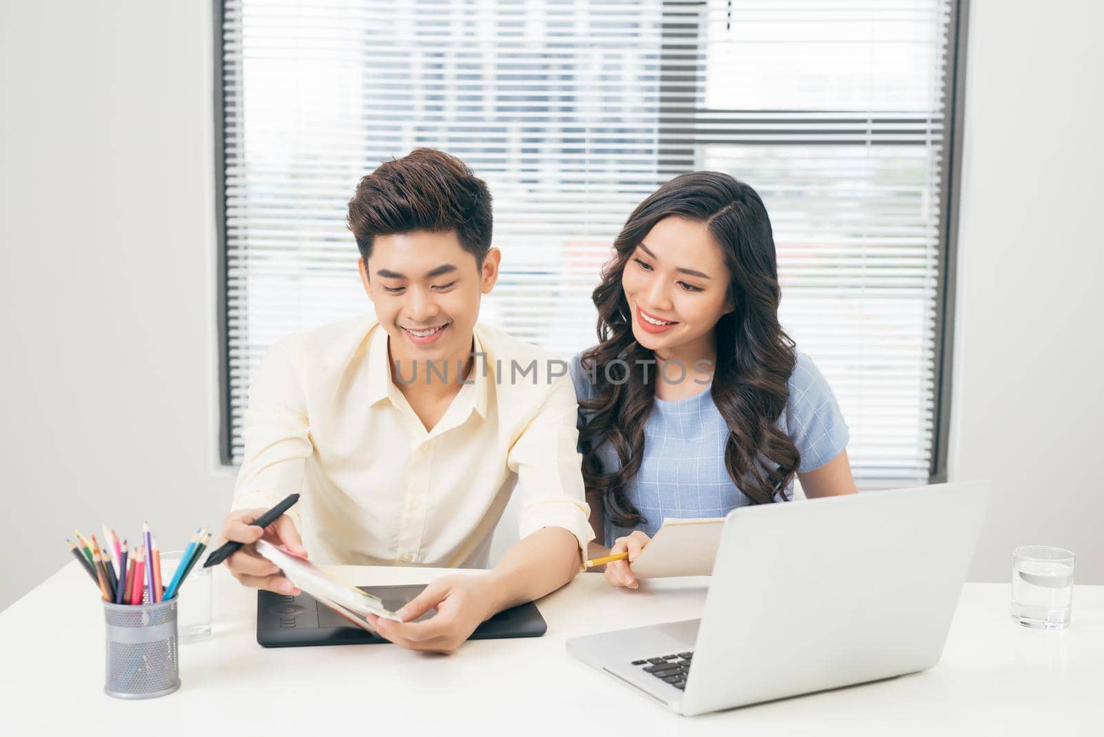 Two smiling casual designers working with laptop while sitting at desk in the office