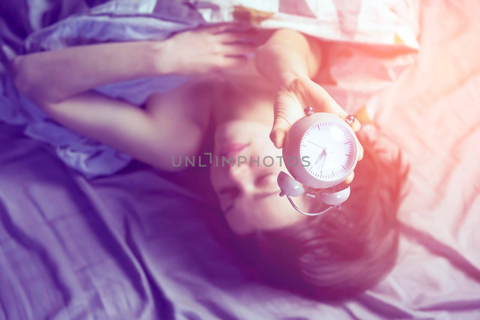 A young girl sleeps early in the morning under the blanket and holds a vintage alarm clock in her hands.