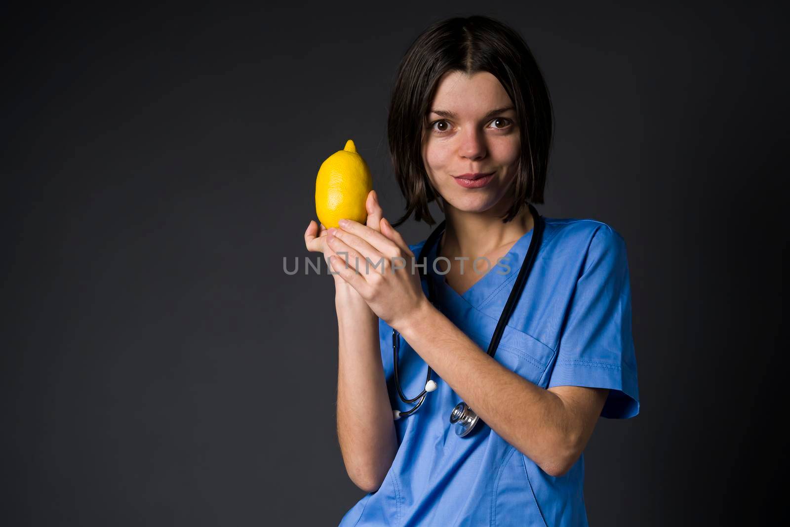 A young girl cheerful doctor in a blue medical suit and with a stethoscope holds a lemon fruit in her hands and smiles. The nurse is holding a nutritious fruit containing vitamin C.