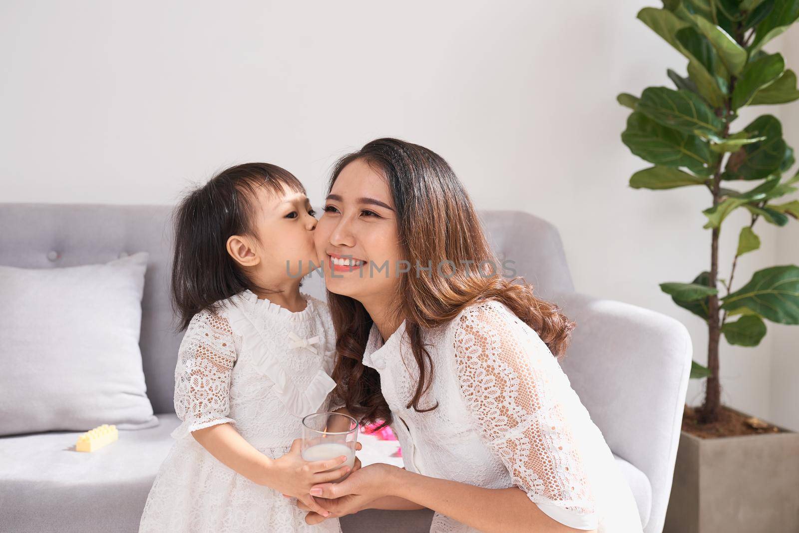 Little girl and her mom drinking milk sitting on sofa at home. Motherhood and care, healthy eating and lifestyle, early development concept, copy space