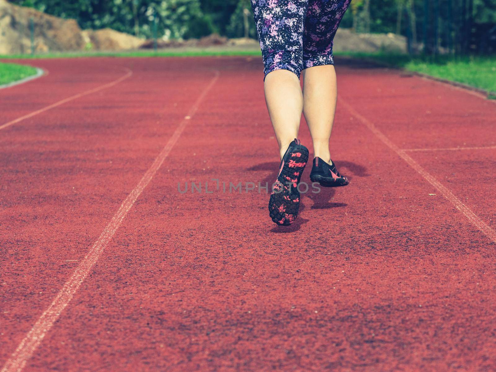 Mature asian woman in flowered leggings up her knees and toe shoes running along the stadium track.