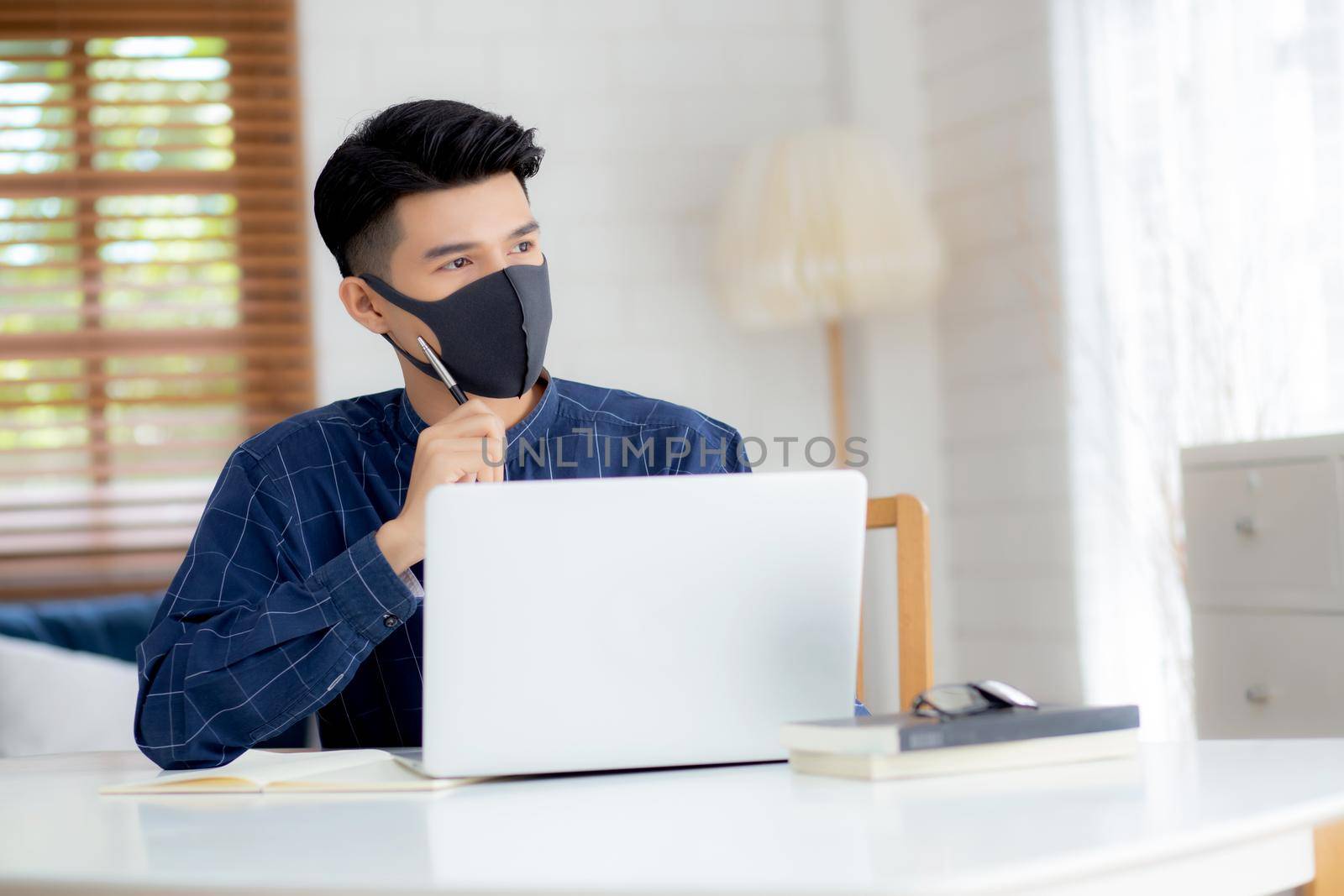 Young business man in face mask planning and working from home on laptop computer on desk for protection for outbreak of covid-19, male quarantine stay home using notebook on desk, new normal. by nnudoo