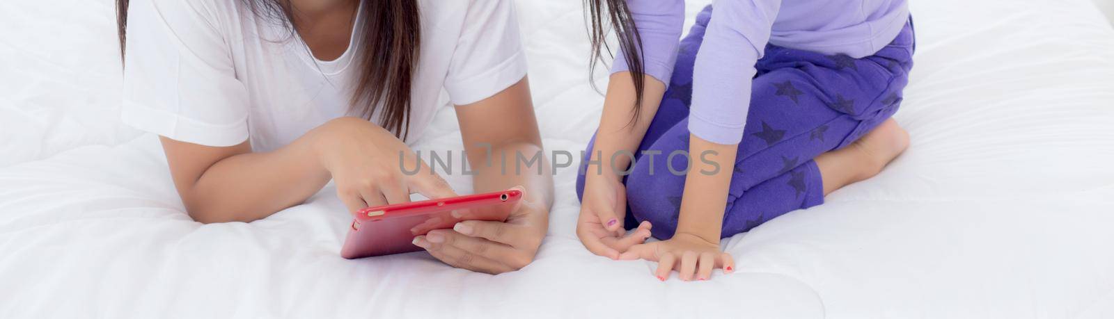 Young asian mother and little daughter looking tablet computer together for learning and education on bed in bedroom, mom and girl watching tablet to internet at home, family and technology concept. by nnudoo