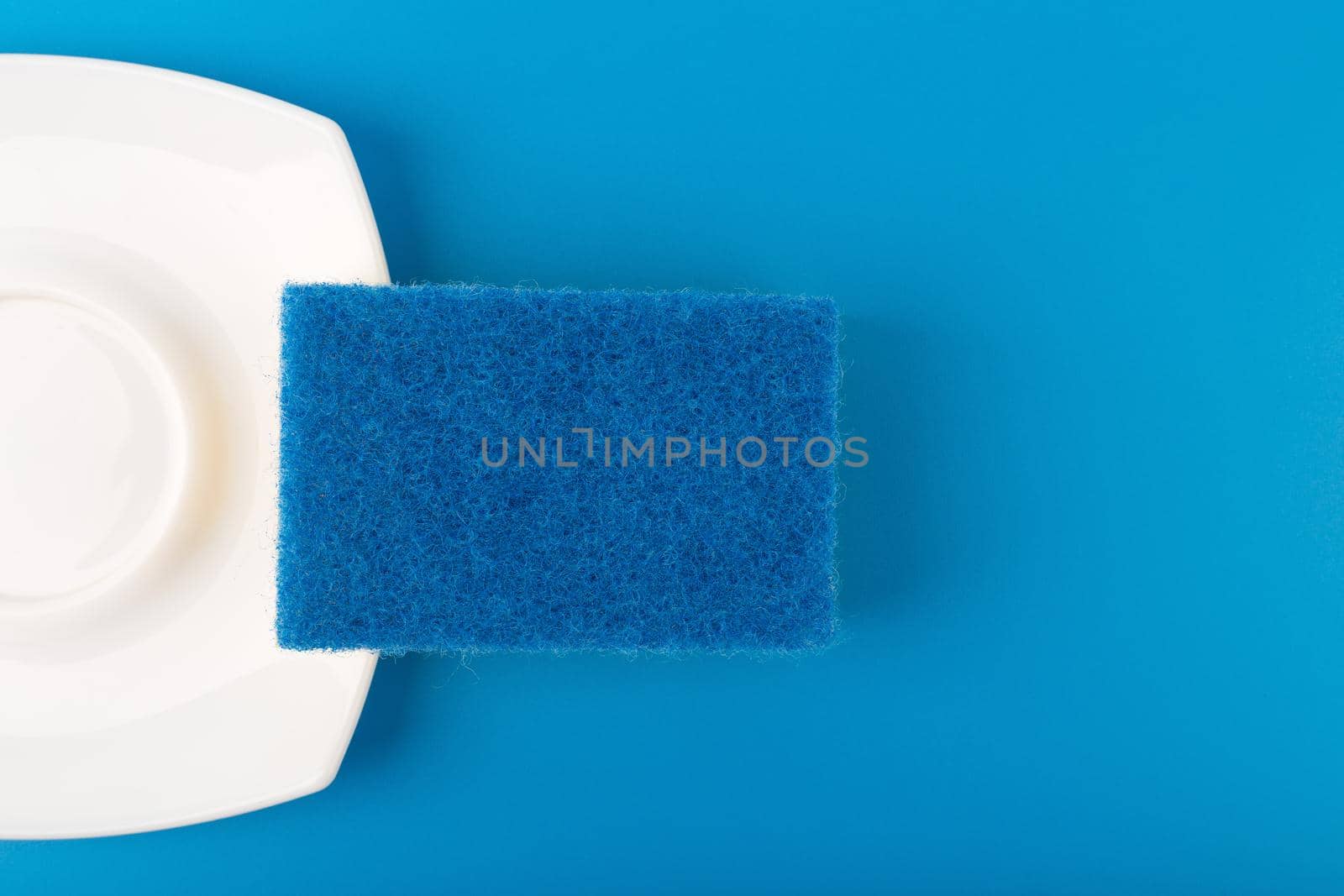 Dishwashing minimal creative concept. Close up of white shiny clean plate and blue cleaning sponge against blue background with copy space.