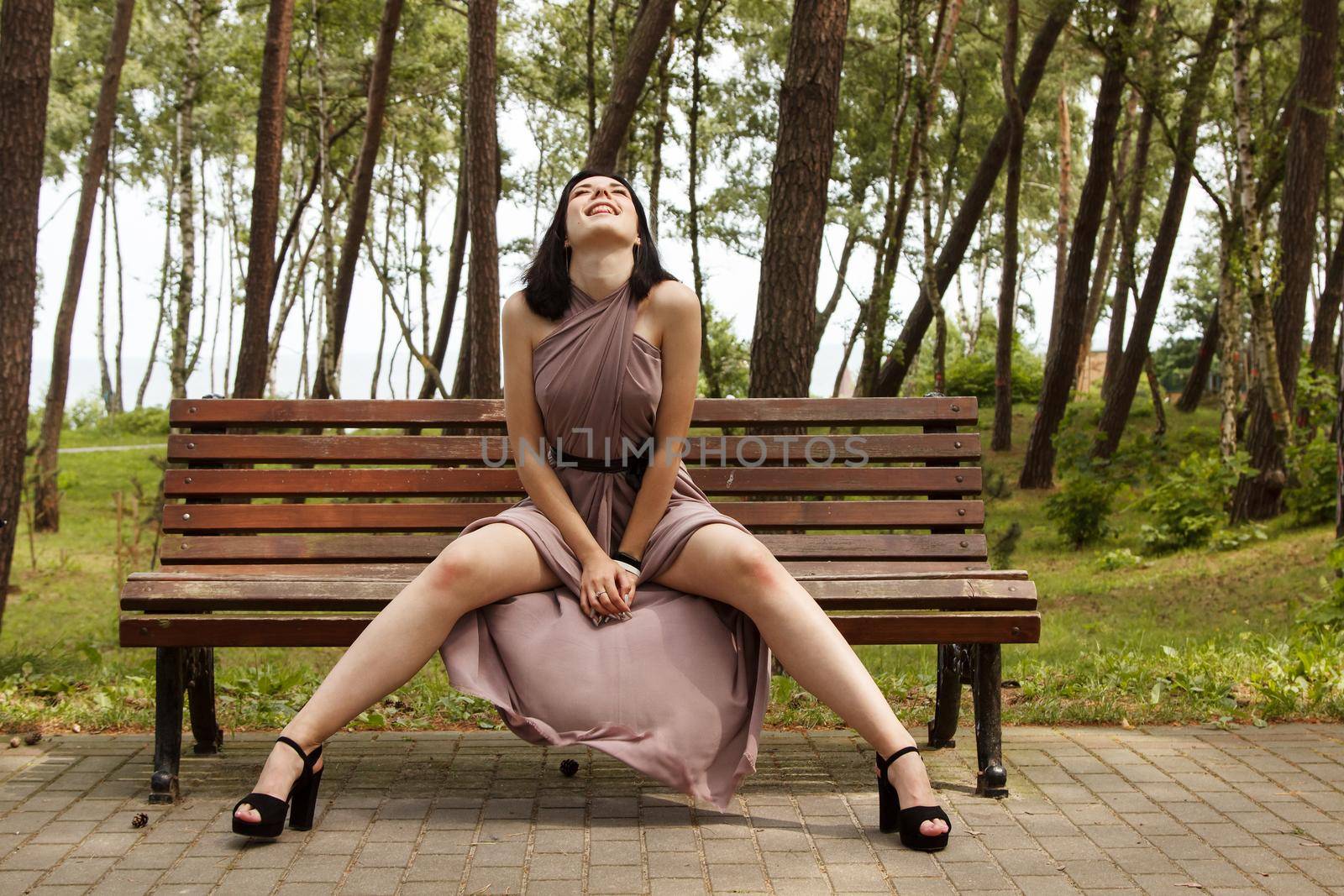 young beautiful brunette woman in beige dress sitting on a park bench on summer day