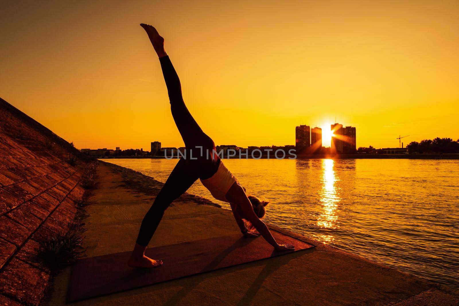 Young woman is exercising outdoor. She is practicing yoga on sunny day. Eka Pada Adho Mukha Svanasana/One-Legged Downward-Facing Dog Pose.