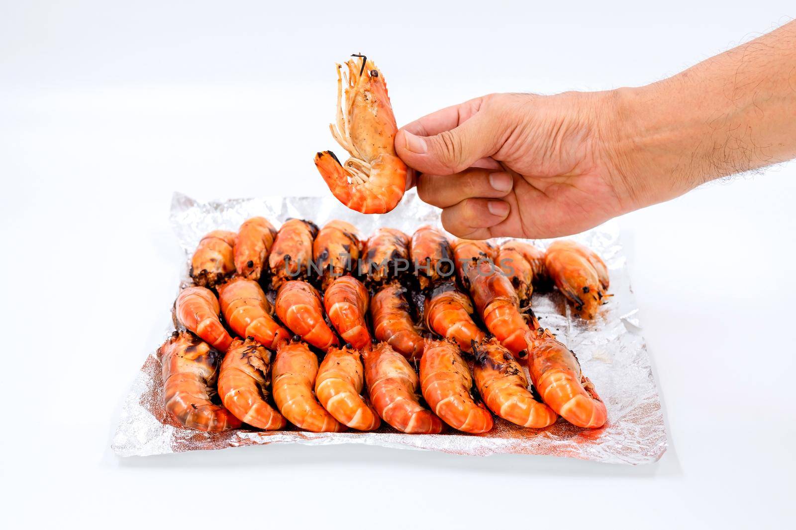 Close up a human hand picks up grilled river prawns from a pile on foil on white background. by wattanaphob