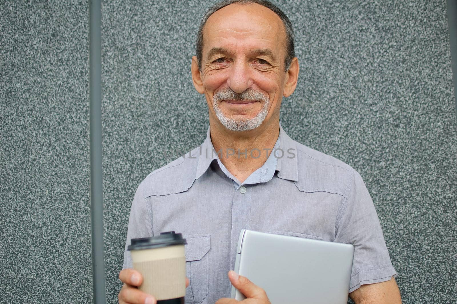 Male portrait of older coworker with silver laptop and cup of coffee waiting for a meeting near dark grey wall in the morning, remote job, coffee break.
