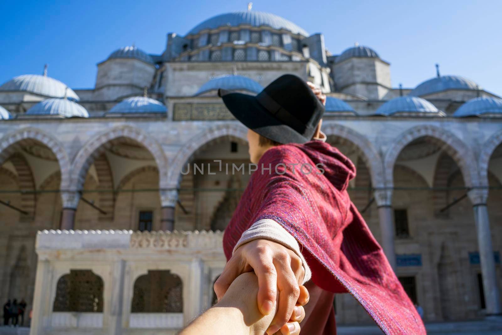 A young traveler girl in a red poncho and hat holds a friend's hand, follow me gesture. A couple in love travels to Arab countries. An ancient blue mosque on the background, Istanbul, Turkey.