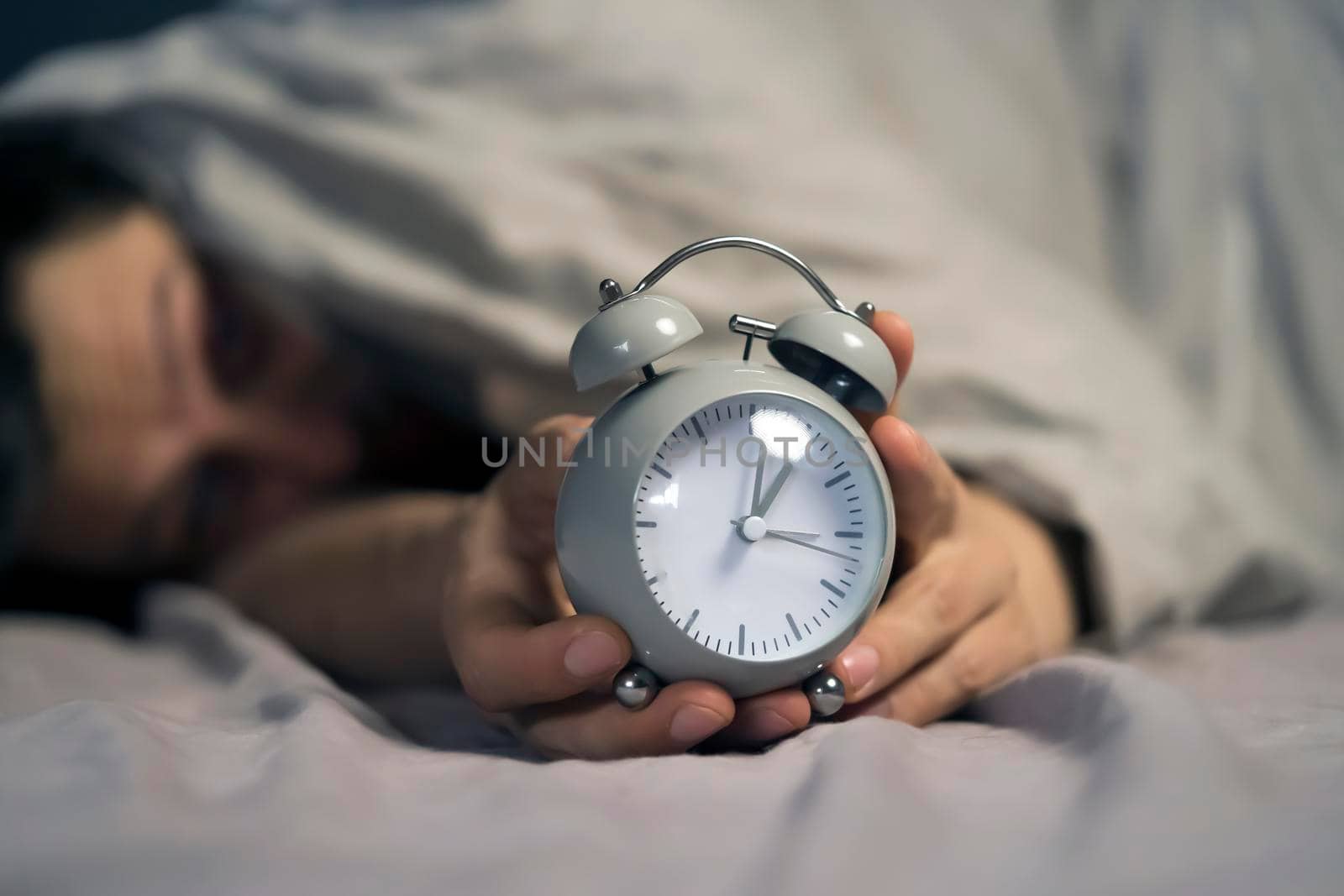 Hands of a young man from under the blankets hold a retro vintage alarm clock. by africapink