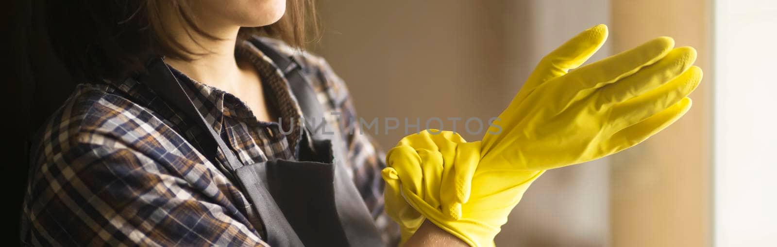 A young girl in a plaid shirt and apron puts yellow rubber gloves on her hands to start cleaning her house and create comfort. Housekeeper with gloves doing disinfection.