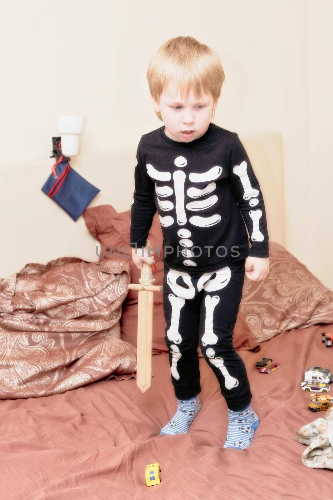 A child in a skeleton costume with a wooden sword plays on the couch. Children games, fears and horror stories