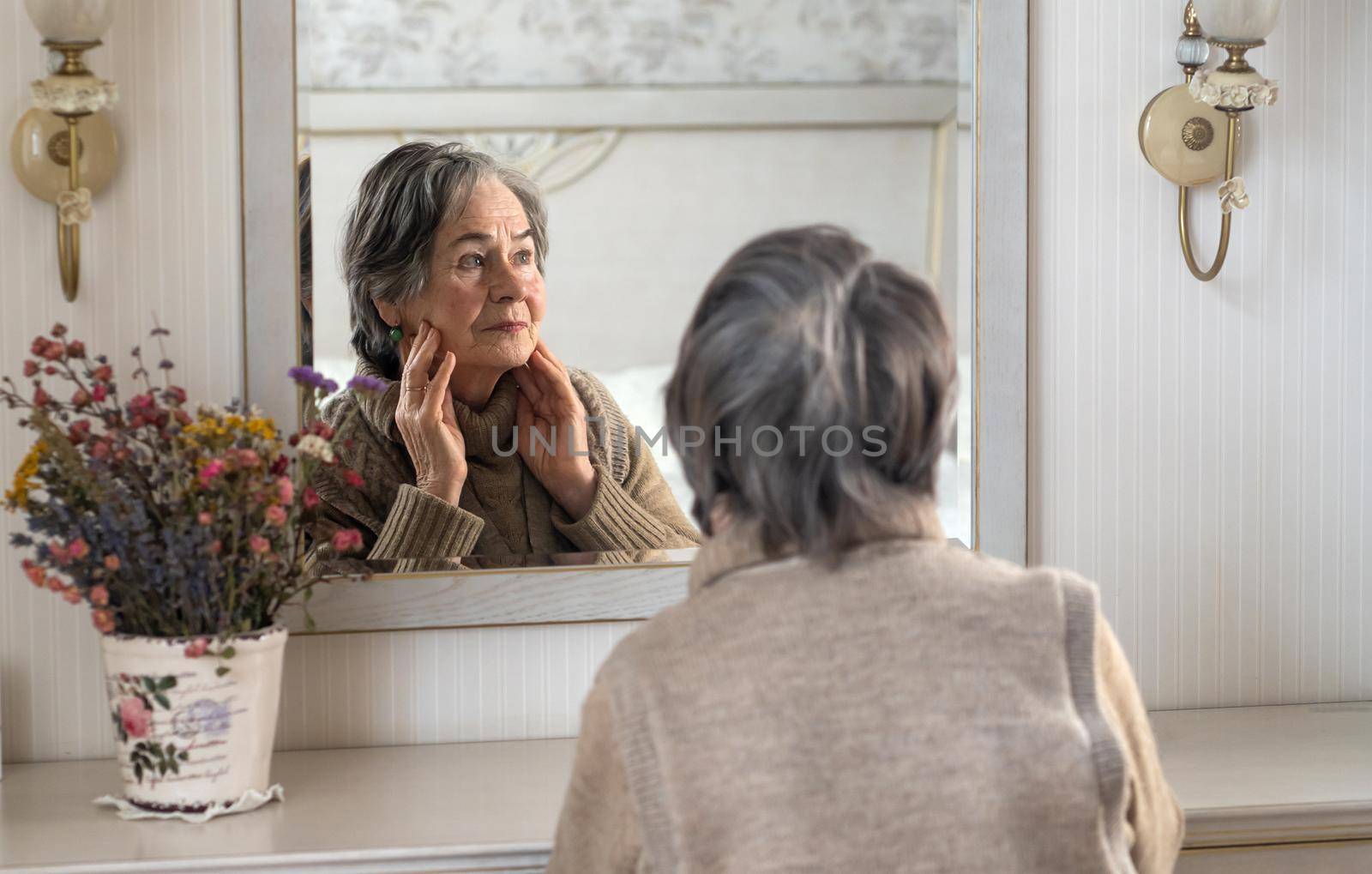 An elderly well-groomed gray-haired woman looks at herself in the mirror while sitting at the cosmetic table. Old lady, pensioner does facial skin care in her cozy home.