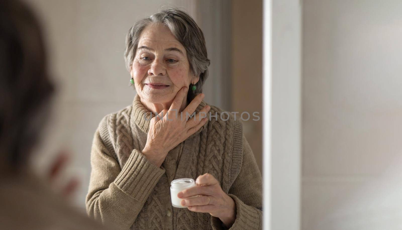 An elderly woman applies an anti-aging moisturizer. The pensioner looks after her appearance, makes cosmetic procedures in the bathroom. Uses cream for skin rejuvenation, regeneration, lifting.