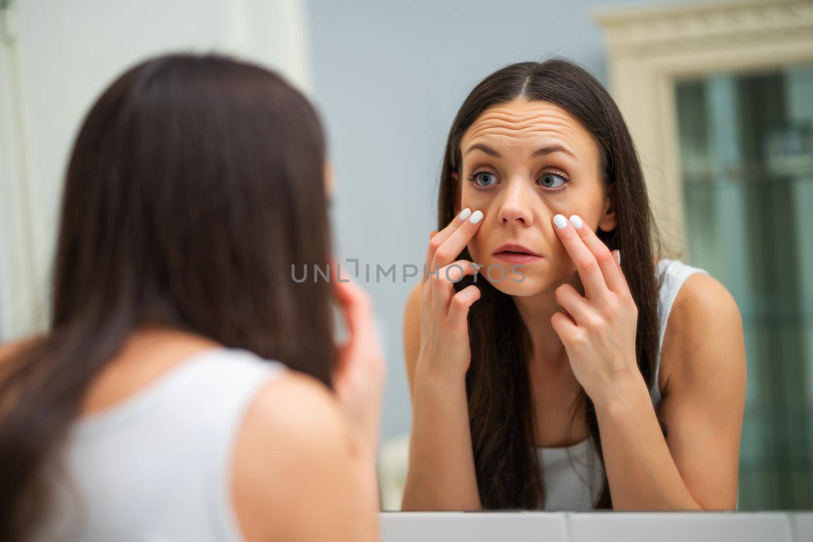 Tired woman looking at her eye bags in the bathroom.