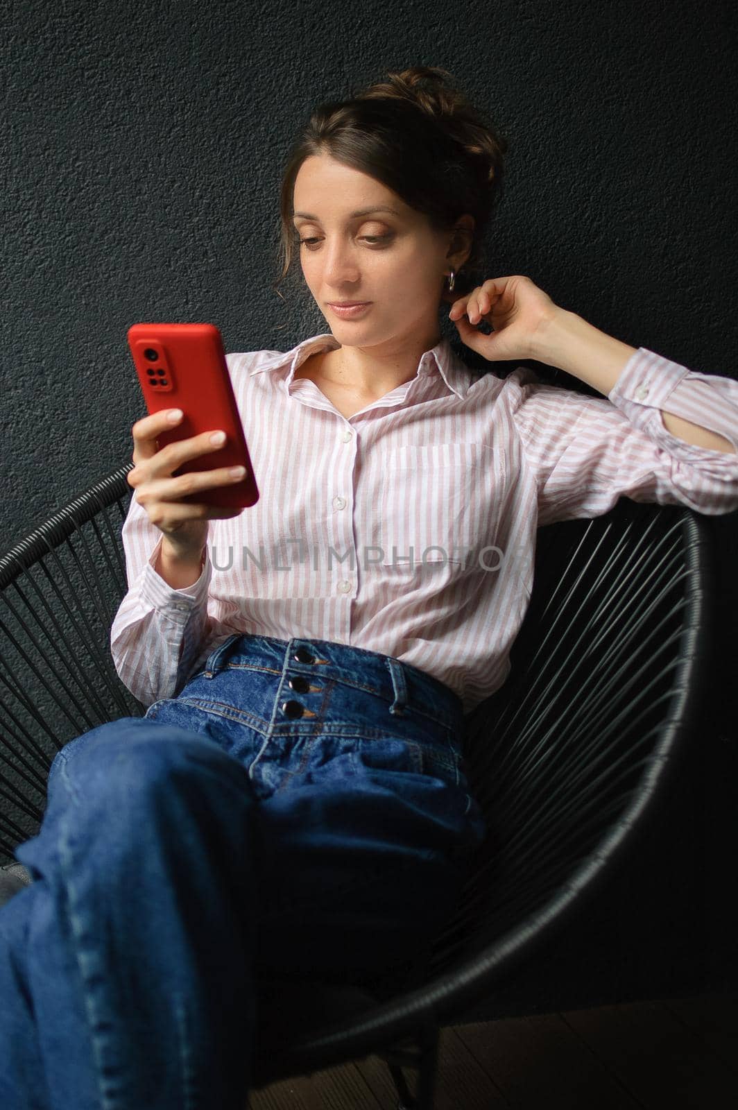 Beautiful curly girl is using red smartphone at home in her apartment, on dark background, happy people concept.