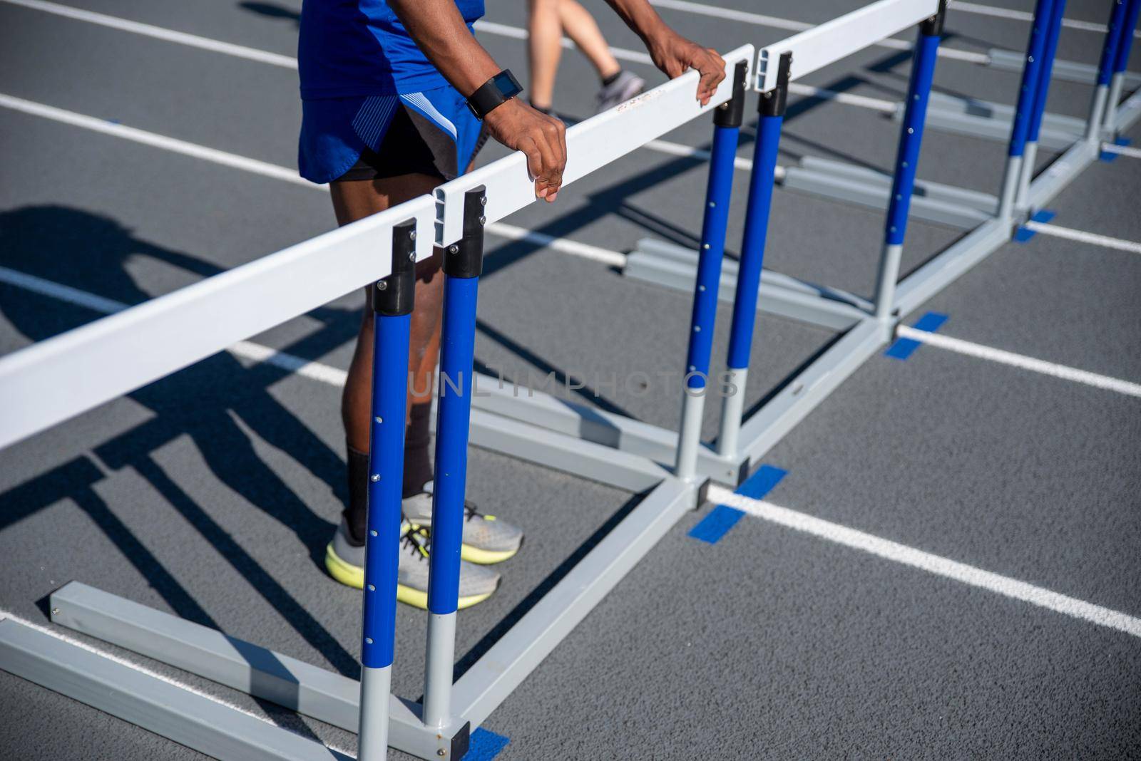 A long row of hurdles blocks the track and one young African American man straightens them. Track is new looking, gray with crisp white lines.