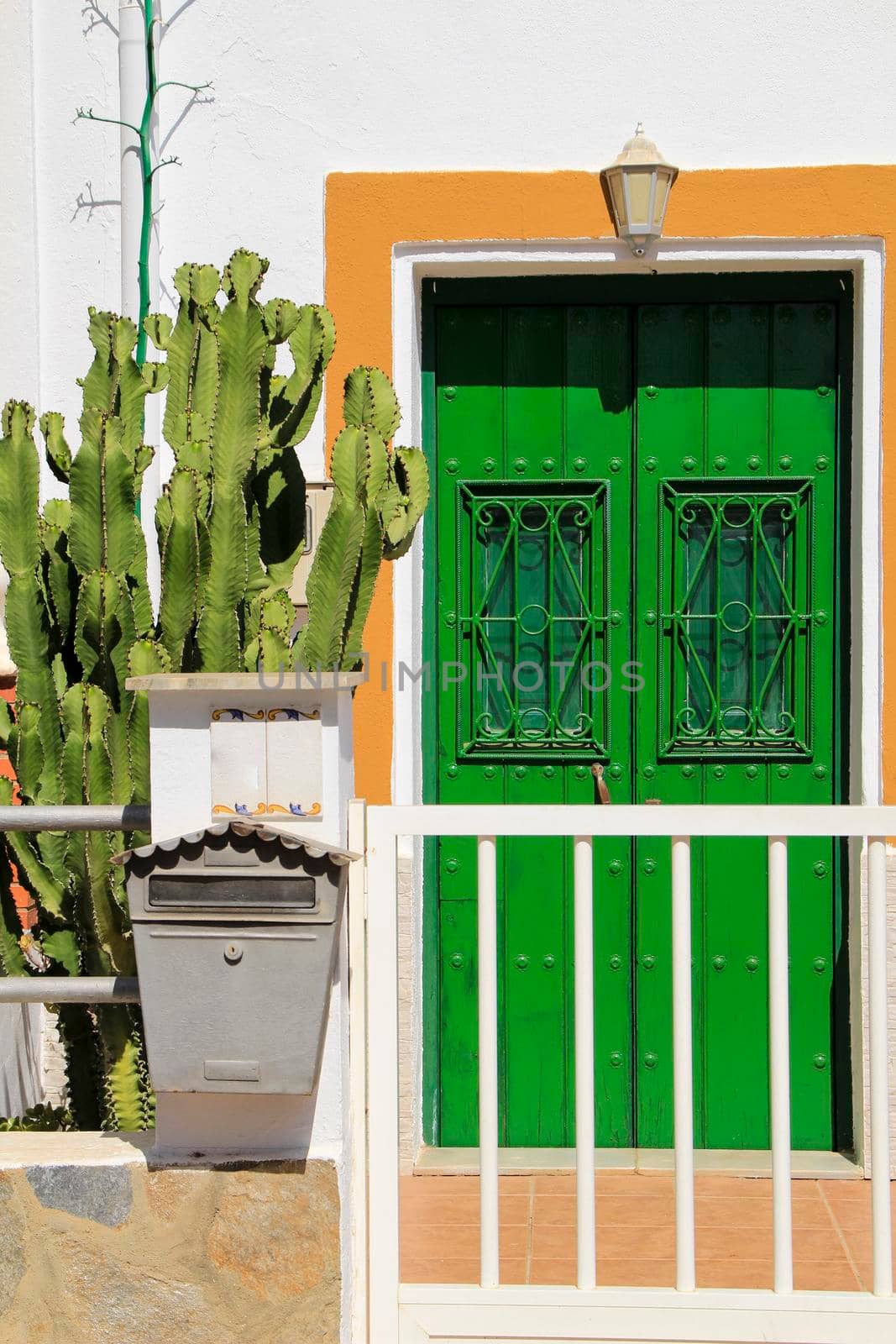 Wooden door painted green with metal details. White fence, mailbox and cactus pots at the door.