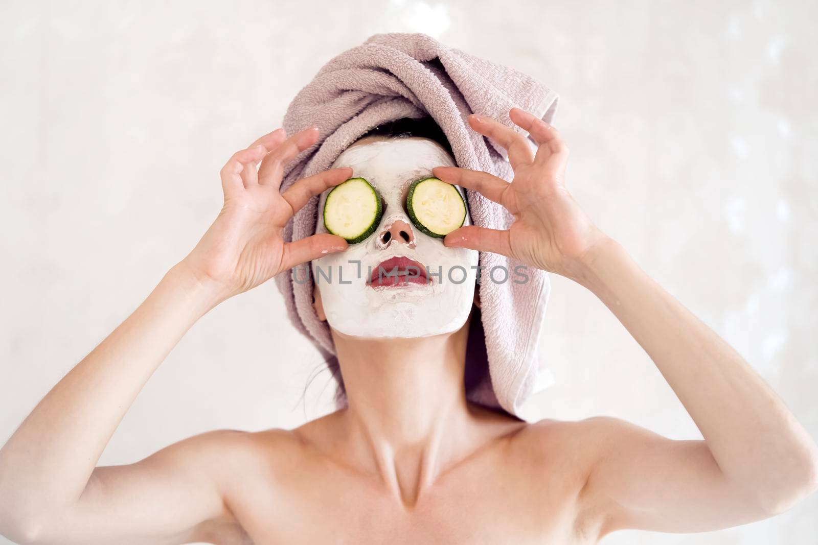 A young girl takes care of the health of her skin, woman applies a mask of white clay to her face and puts cucumbers on her eyes in the bathroom near the mirror.