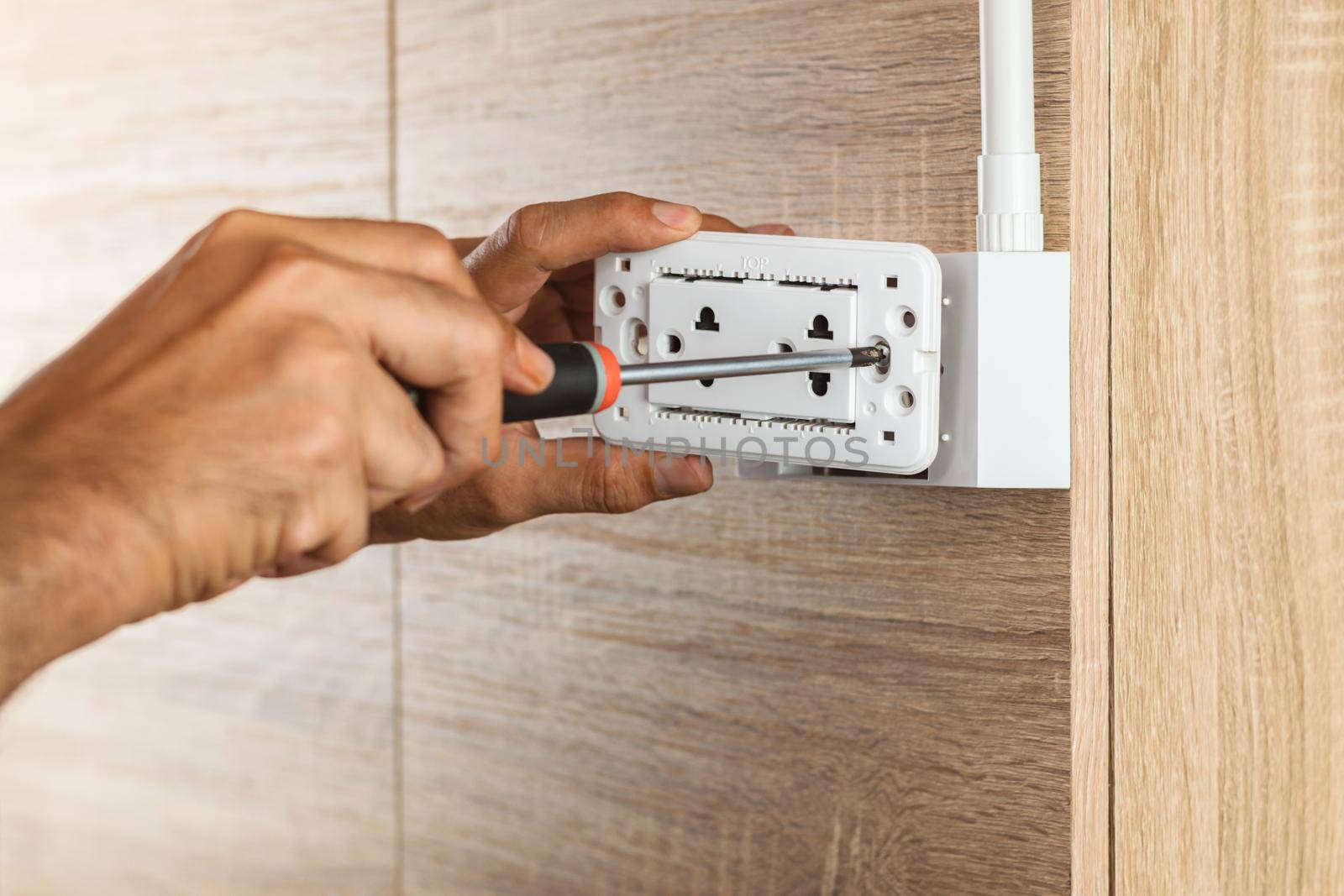 Electrician is using a screwdriver to install the electric power socket in to a plastic outlet box on a wooden wall. by wattanaphob