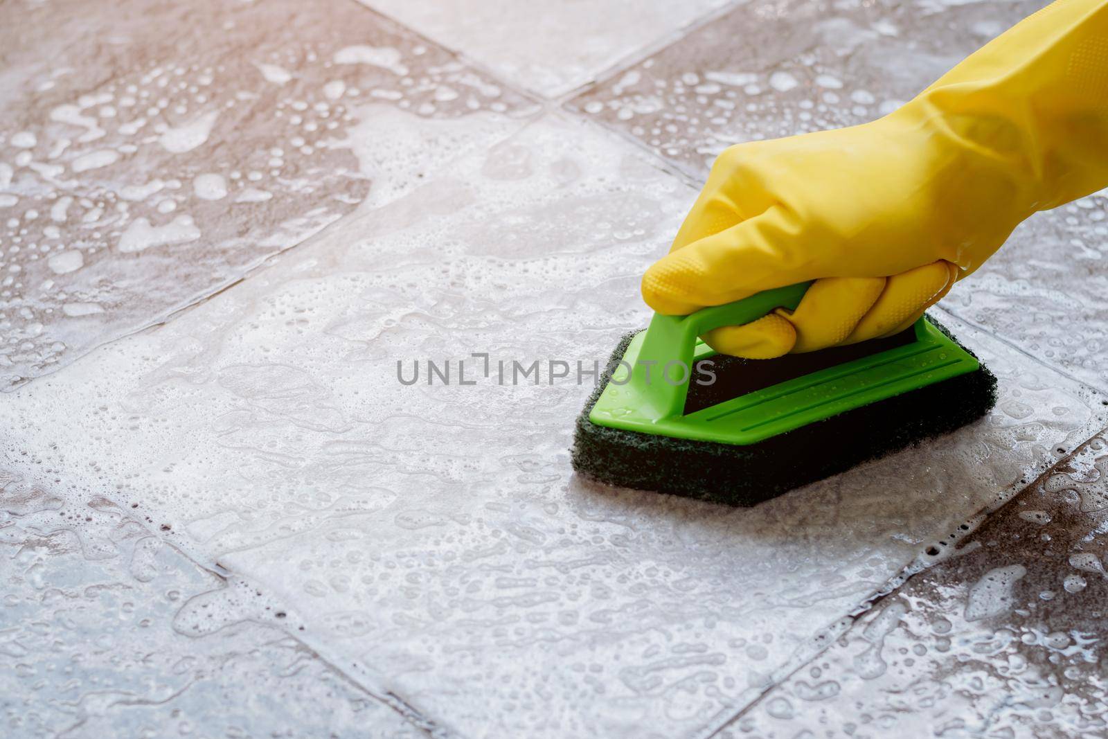 Human hands wearing yellow rubber gloves are using a green color plastic floor scrubber to scrub the tile floor with a floor cleaner. by wattanaphob