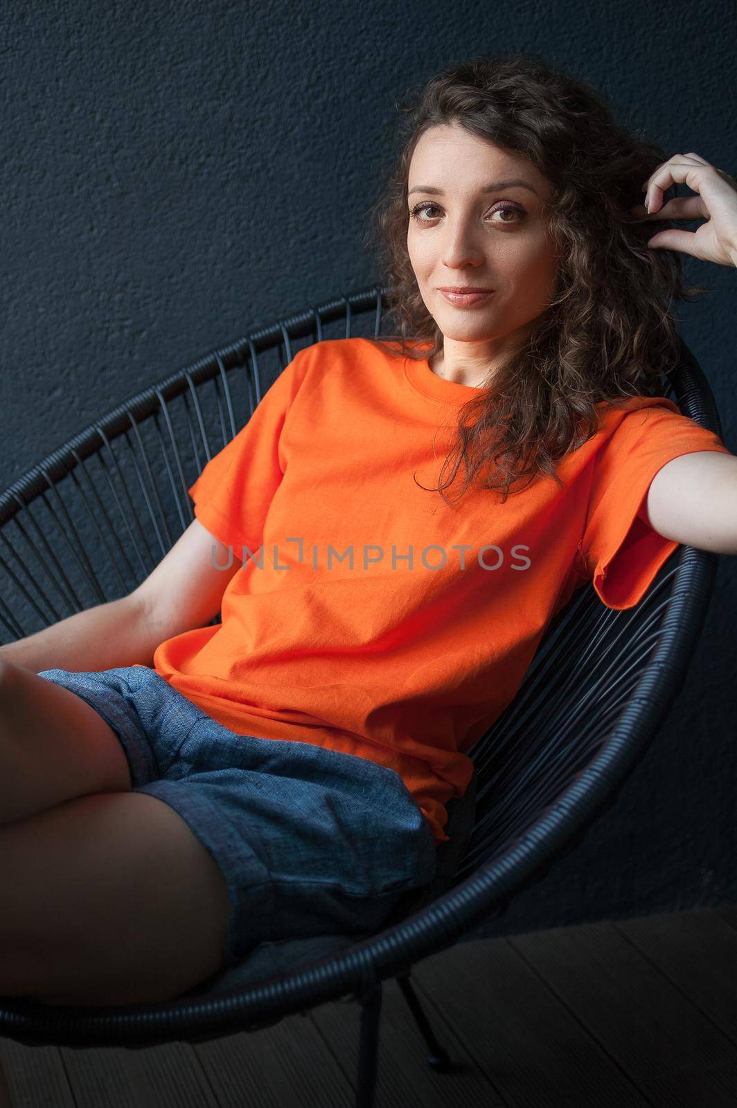 Female portrait of a beautiful curly girl in bright orange t-shirt sitting at home in her apartment on dark background, happy people concept.