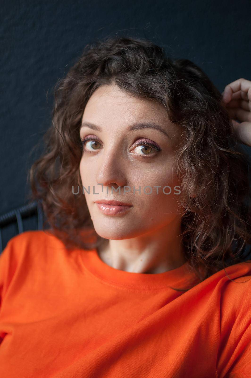 Female portrait of a beautiful curly girl in bright orange t-shirt at home in her apartment on dark background, happy people concept by balinska_lv