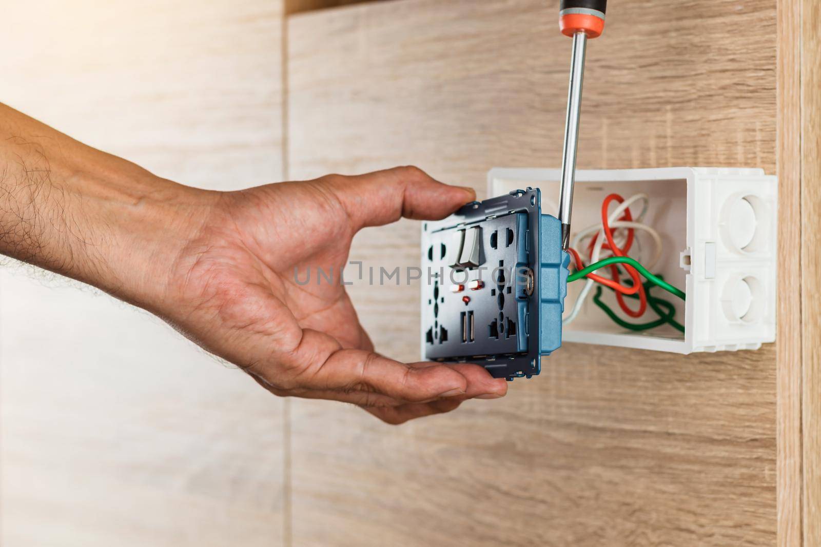 Hand of an electrician is using a screwdriver to attach the wires to the universal wall outlet AC power plug with USB port and on-off in a plastic box on a wooden wall. by wattanaphob