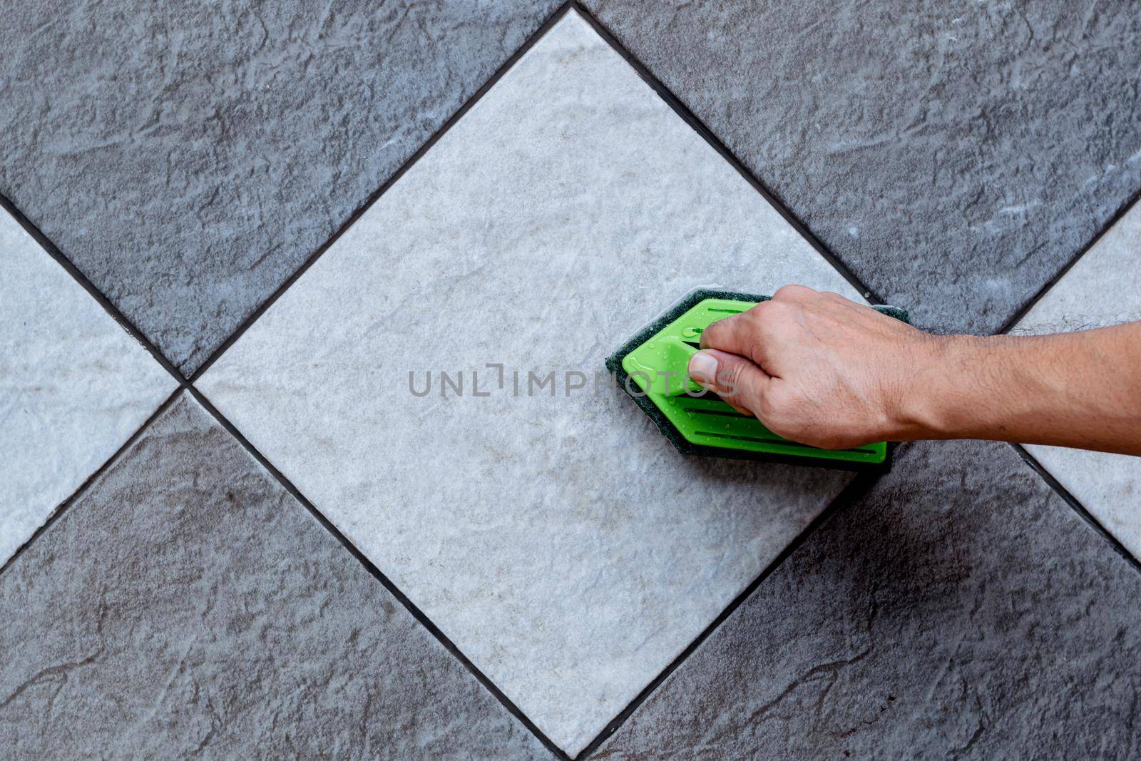 Top view of a human hand are using a green color plastic floor scrubber to scrub the tile floor with a floor cleaner.