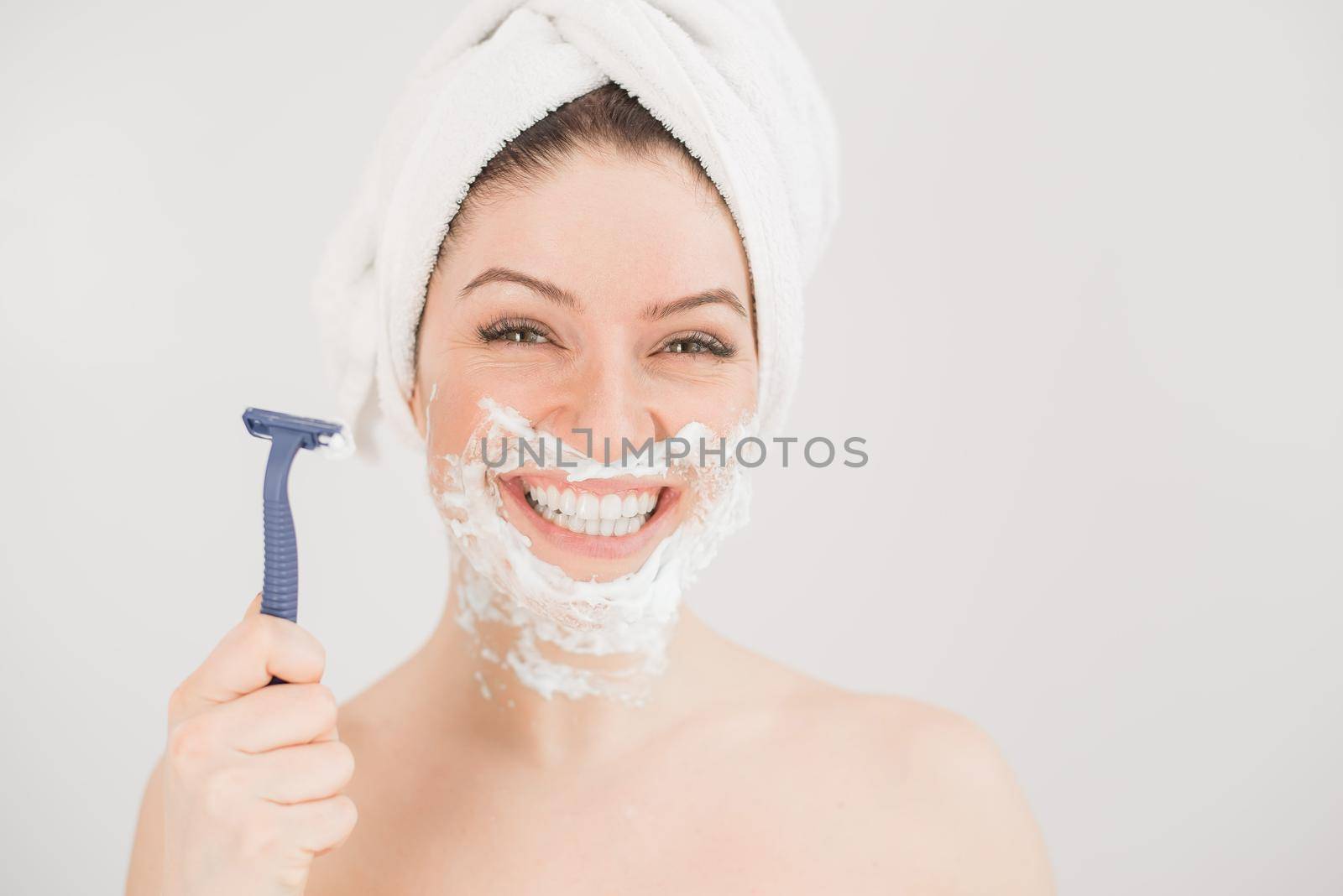 Cheerful caucasian woman with a towel on her head and shaving foam on her face holds a razor on a white background by mrwed54