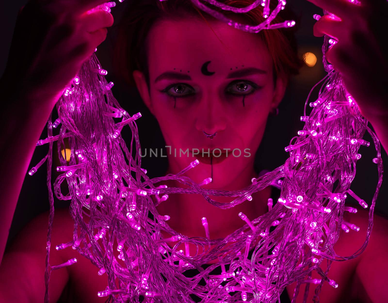 Woman with witch makeup and an earring in her nose. The girl holds a garland of flickering pink lights near her face. Light bulbs illuminate the witch's face in the dark. by mrwed54