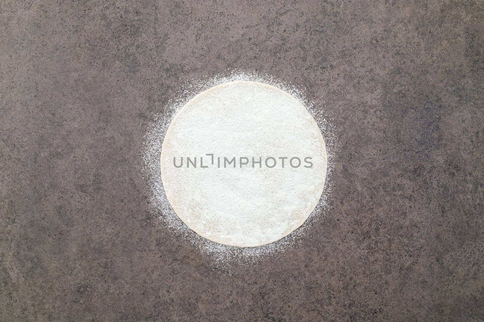 Thin homemade pizza dough with scattered wheat flour on dark concrete background. by kerdkanno