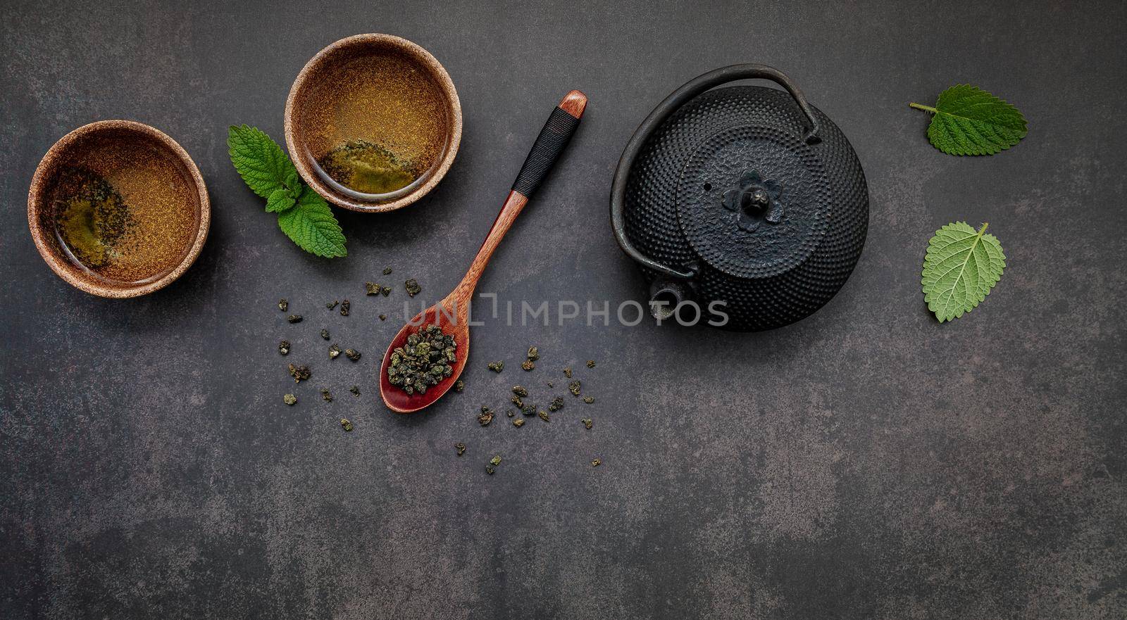 Black cast iron tea pot with herbal tea set up on dark stone background.