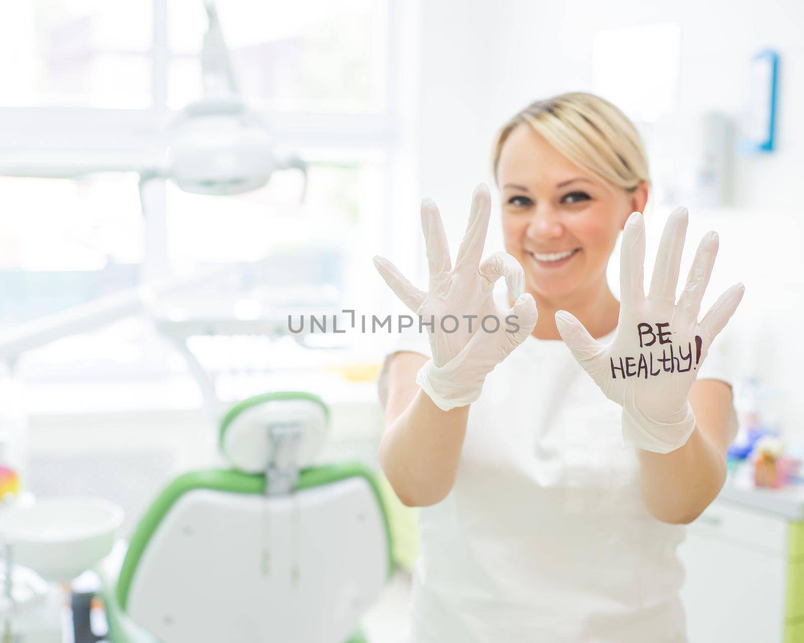 Friendly female dentist smiling and showing okay sign with fingers. Blonde doctor in gloves in the office in the medical center. The inscription on the hand be healthy.