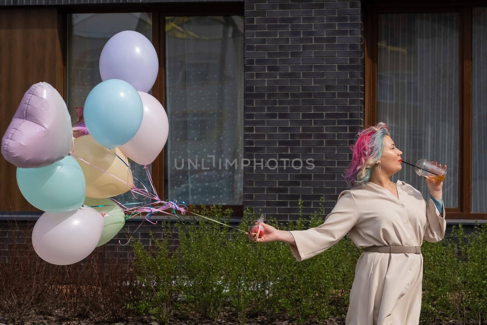 Woman in colored hair walks with an armful of balloons and drinks a refreshing beverage.