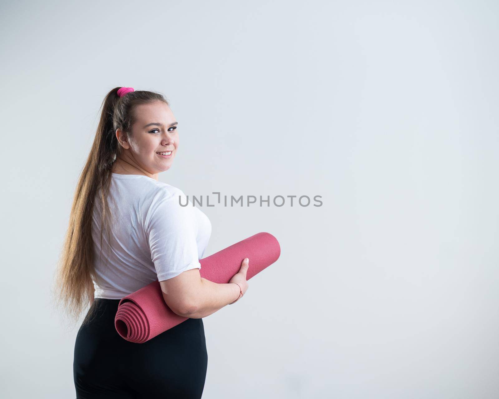 Young fat caucasian woman holding a sport mat. Charming plus size model in sportswear stands on a white background by mrwed54