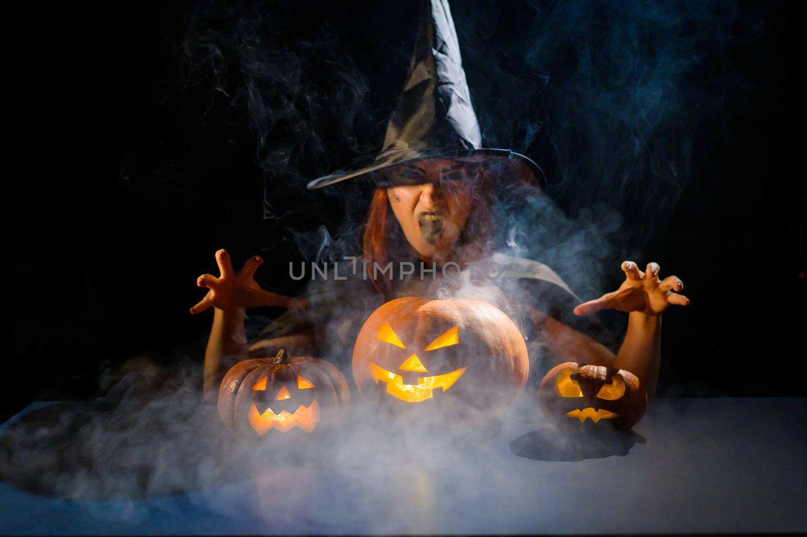 The evil witch casts a spell on pumpkins. Portrait of a woman in a carnival halloween costume in the dark by mrwed54