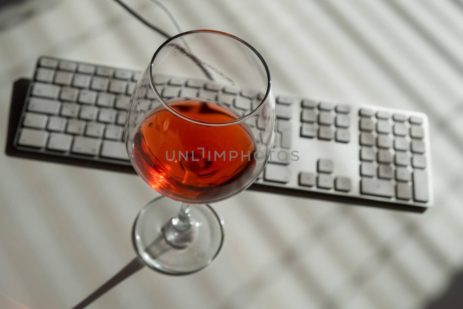 A goblet with red wine and a keyboard on a white table with shade from blinds.