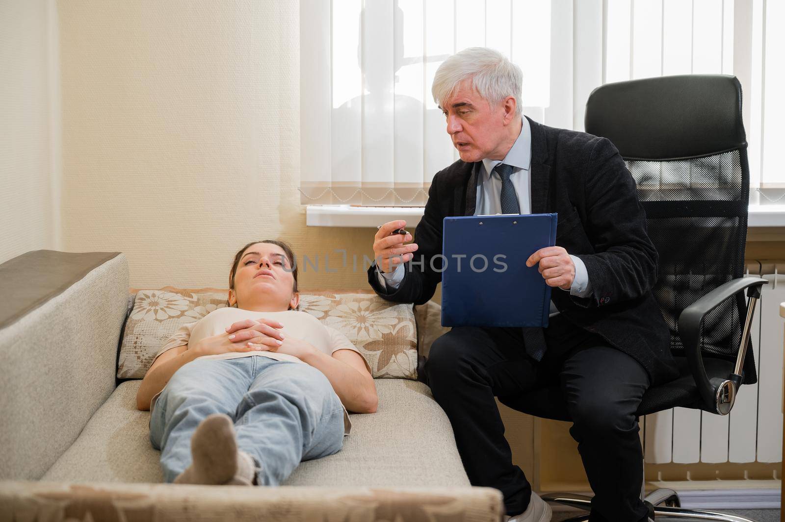 Caucasian woman lies on a couch at a reception with a psychotherapist. An elderly man works as a psychiatrist.