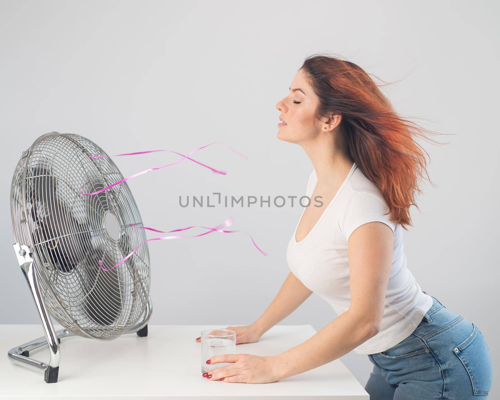 A red-haired Caucasian woman chills by the electric fan and drinks a cold drink. Climate control in the apartment.