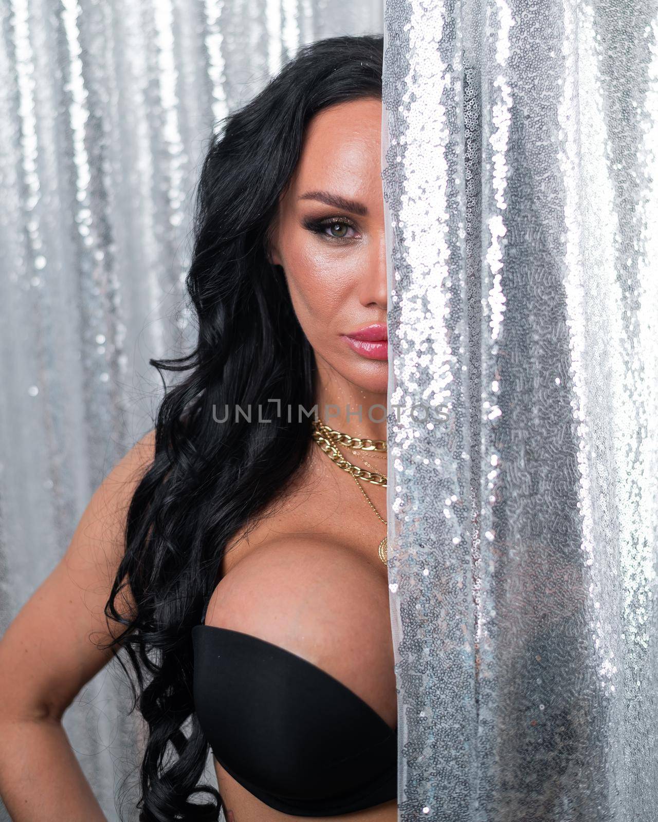 Portrait of a sexy brunette woman with big breasts dressed in a black bra peeking out from behind curtains of silver sequins