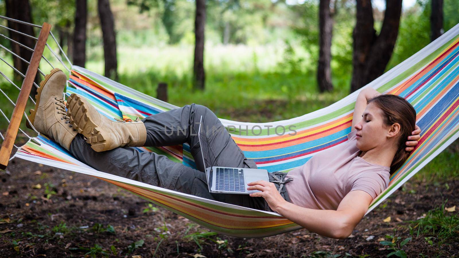 Caucasian woman working on laptop while sitting in a hammock in the forest. Girl uses a wireless computer on a hike