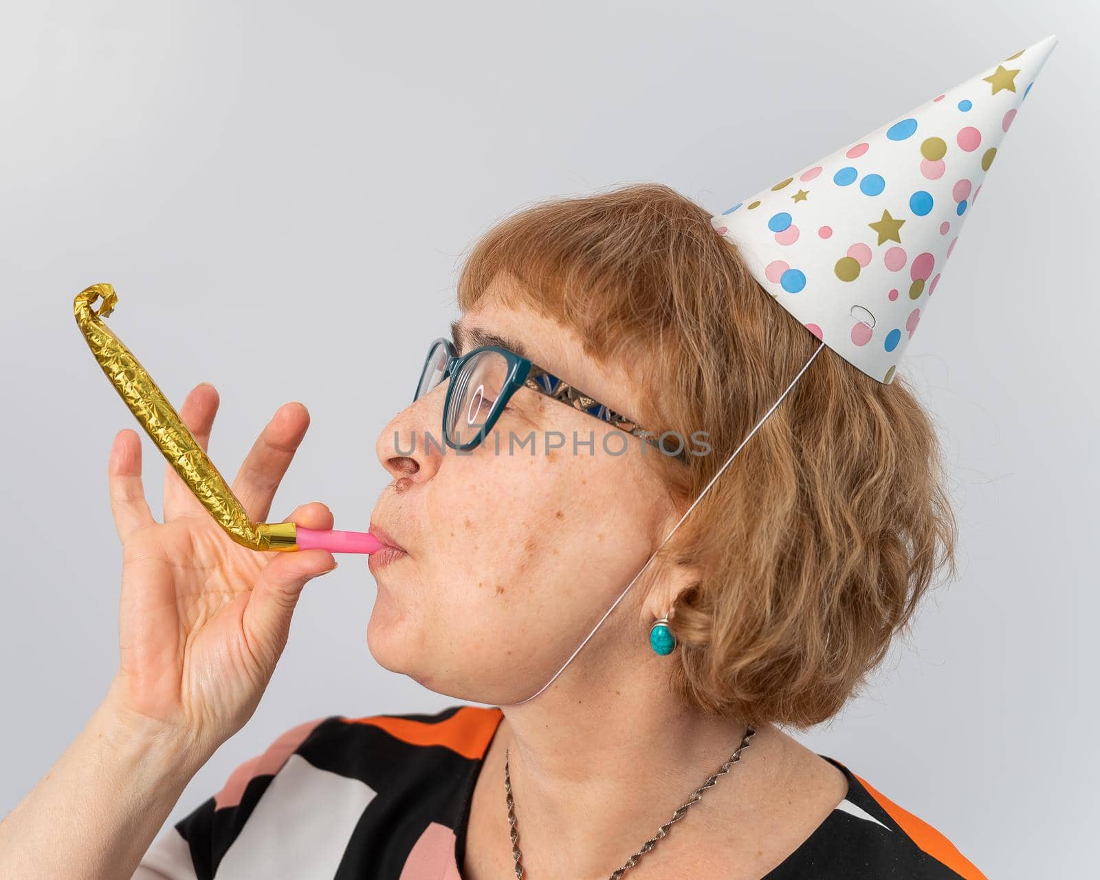 Portrait of a smiling elderly woman in a festive cap holding a whistle tongue on a white background.