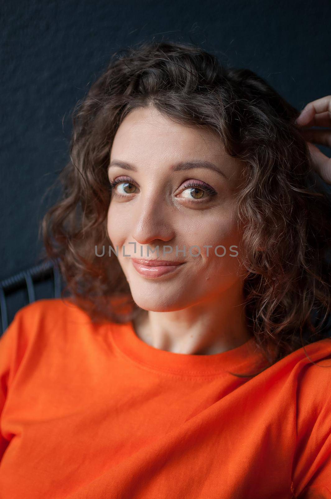 Female portrait of a beautiful curly girl in bright orange t-shirt at home in her apartment on dark background, happy people concept.