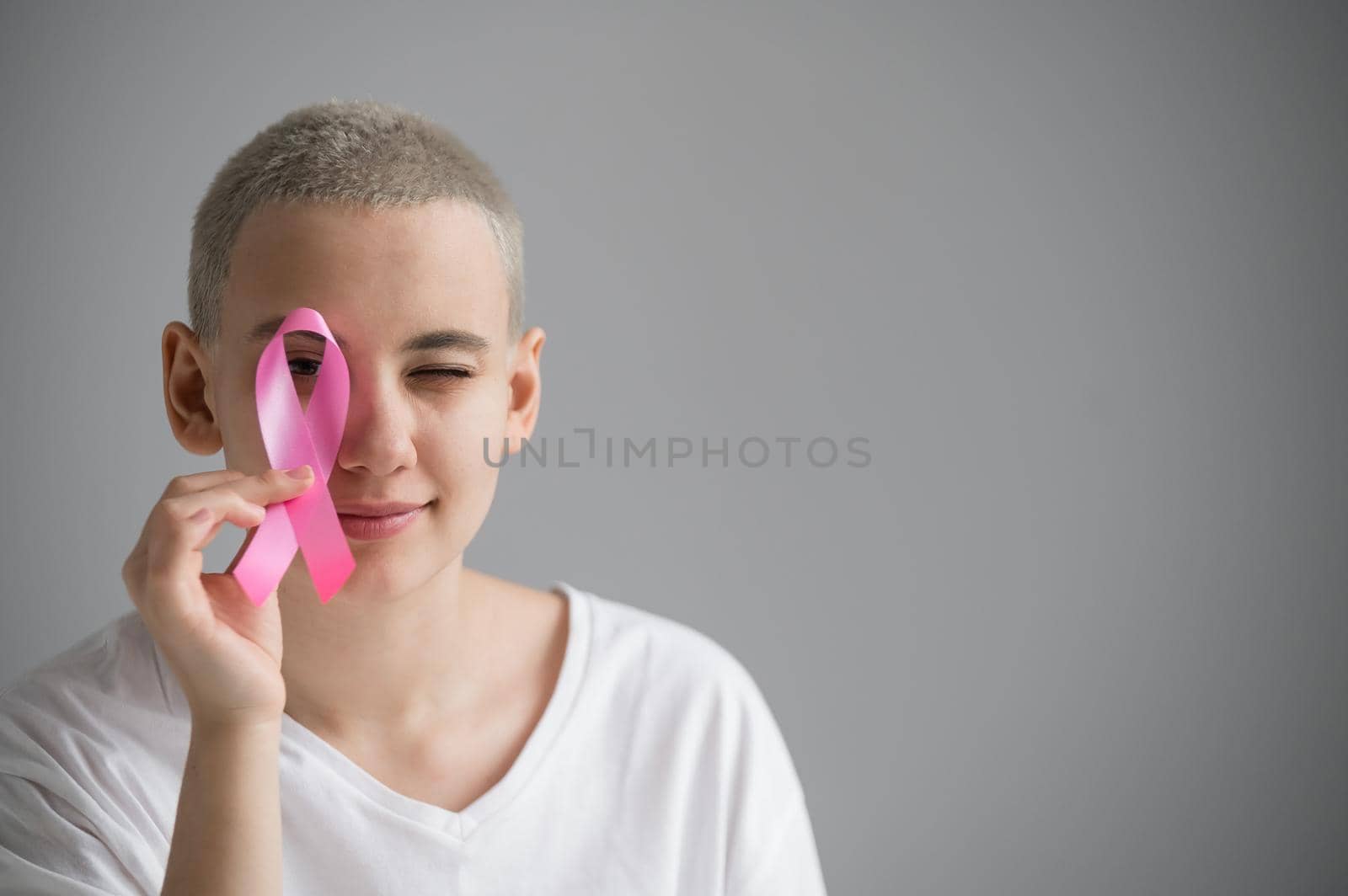 Young woman with short hair wearing a white t-shirt holding a pink ribbon as a symbol of breast cancer on a white background